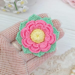 Puede incluir: Una flor de ganchillo con un centro amarillo brillante, múltiples capas de pétalos rosas y una base de hojas de color verde claro. El diseño floral está elaborado con hilo fino, mostrando intrincados patrones de puntos y textura.