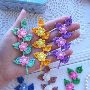May include: Three crochet flower and leaf appliques in mint green with pink flowers, yellow with yellow flowers, and purple with light purple flowers. Each flower features a small white pearl bead at the centre. The stems and leaves are textured.