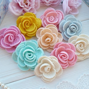 Puede incluir: Una colección de nueve flores de ganchillo en varios colores, incluyendo amarillo, rosa claro, azul claro, melocotón, verde menta y crema. Cada flor presenta pétalos redondeados y superpuestos en forma de rosa abierta.