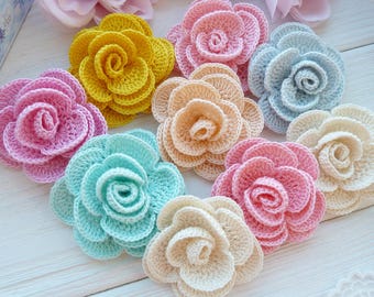 May include: A collection of nine crocheted flowers in various colors including yellow, light pink, light blue, peach, mint green, and cream. Each flower features layered, rounded petals arranged in a circular, blooming rose-like pattern.