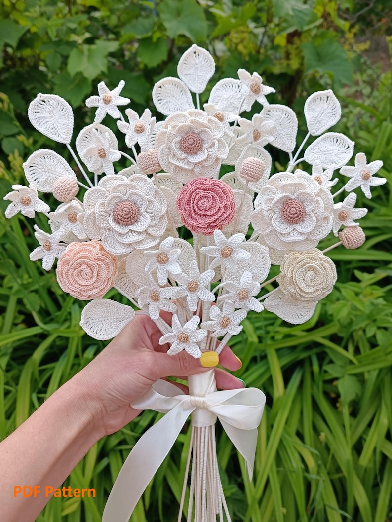 Puede incluir: Un ramo de flores de ganchillo hecho a mano con flores blancas, crema y rosas. El ramo est&aacute; atado con una cinta de raso blanca. El texto "PDF Pattern" es visible en la parte inferior. Las flores est&aacute;n hechas con intrincados detalles de ganchillo.