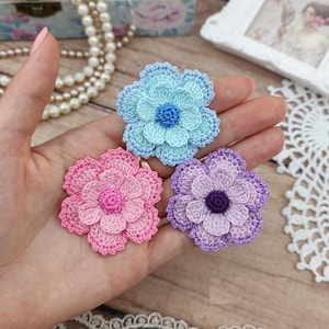 Puede incluir: Tres aplicaciones de flores de ganchillo en azul claro, rosa y morado. Cada flor presenta pétalos en capas y un centro de color sólido que contrasta. La flor azul claro tiene un centro azul, la flor rosa tiene un centro rosa y la flor morada tiene un centro morado oscuro.