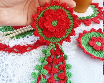 May include: A round, crocheted ornament featuring a central red flower with a green center, surrounded by a green border with a scalloped edge. Three dangling strands of small red berries and green leaves hang from the bottom of the ornament.