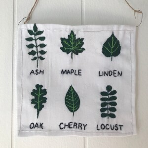 May include: A white fabric wall hanging with six embroidered green leaves and their corresponding names: Ash, Maple, Linden, Oak, Cherry, and Locust.