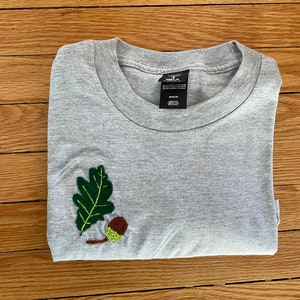 May include: Gray t-shirt with an embroidered green oak leaf and brown acorn on the left side. The shirt is folded in half and the tag reads "Medium".