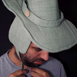 May include: A light green hat with a wide brim and a conical crown. The hat features a strap with a wooden button and ear flaps. The hat is made of a textured fabric and is likely a costume or novelty item.
