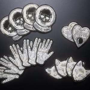May include: A set of iron-on patches featuring various occult and mystical designs. The patches include a skull in a sphere, a hand with palmistry lines, a heart with the text "As Above So Below", and a cat with a third eye.