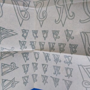 May include: A sheet of paper with a pattern of letters in a triangle shape. The letters are in a cursive font and are outlined in black.
