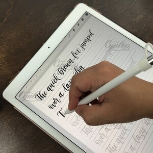 K&ouml;nnte beinhalten: Eine Hand, die einen wei&szlig;en Stift verwendet, um Kalligraphie auf einem digitalen Tablet zu schreiben. Der Text auf dem Bildschirm lautet "The quick brown fox jumped over a lazy dog."