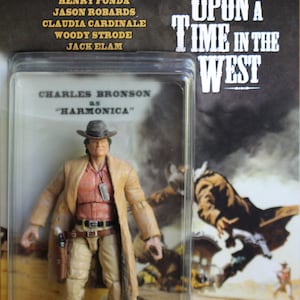 May include: A plastic action figure of Charles Bronson as "Harmonica" from the 1968 Sergio Leone film "Once Upon a Time in the West". The figure is wearing a brown coat, red shirt, and tan pants. The figure is holding a gun in its right hand. The figure is standing on a brown base. The background is a poster of the film.
