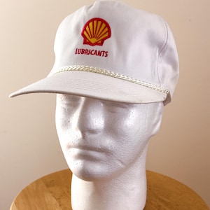 May include: White baseball cap featuring the Shell logo in red and yellow, with the word "LUBRICANTS" embroidered below. The cap has a curved brim and a white rope detail along the edge.