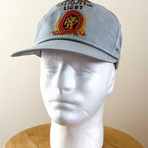 May include: A light gray baseball cap with the text "STROH'S LIGHT" embroidered on the front. The cap features a logo with a lion and wheat stalks in red, gold, and black. A white rope trim accents the brim.