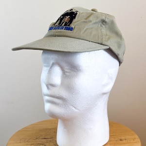 Op de afbeelding: Een kaki baseballpet met een gebogen klep. De pet heeft een geborduurd ontwerp met de tekst "AND LURIN 2000" in blauw en een afbeelding van mensen. De pet wordt getoond op een witte mannequinhoofd.