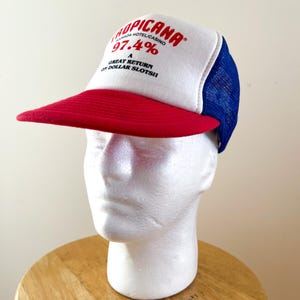 May include: A white, red, and blue trucker hat with the text "Tropicana" and "97.4% Great Return on Dollar Slots!!" printed in red. The hat features a red peak and a blue mesh back.