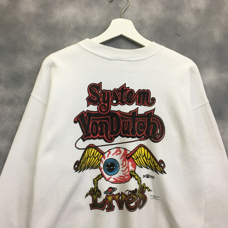 May include: White sweatshirt with a graphic print of a winged eyeball with the text "System Von Dutch Lives" in black and red lettering.