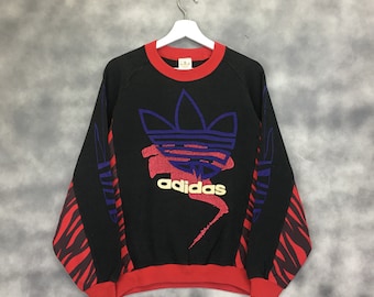 May include: Black sweatshirt with a red and black zebra print on the sleeves. The front features a large, colorful Adidas logo with the word "adidas" in gold lettering.