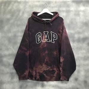 Puede incluir: Una sudadera con capucha de color tie-dye negro y marrón con un logotipo "GAP" blanco en la parte delantera.