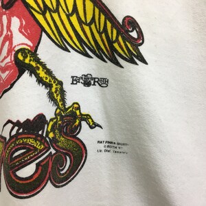 May include: White t-shirt with a red, yellow, and black graphic of a bird with the text "Rat Fink Shirts" and "Roth '93" printed below the graphic.
