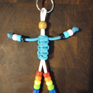 May include: A blue paracord keychain with a wooden bead at the top and rainbow colored beads on the bottom. The keychain is tied in a knot to resemble a person.
