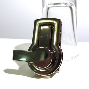May include: A silver-toned metal clasp with a round, rotating knob and a lever handle. The clasp is designed for attaching to bags, purses, or other accessories.