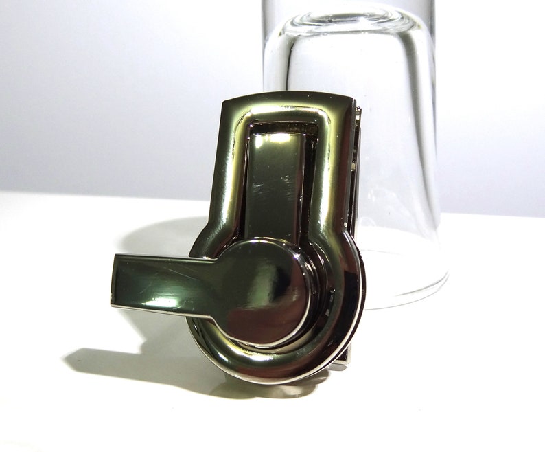 May include: A silver-toned metal clasp with a round, rotating knob and a lever handle. The clasp is designed for attaching to bags, purses, or other accessories.
