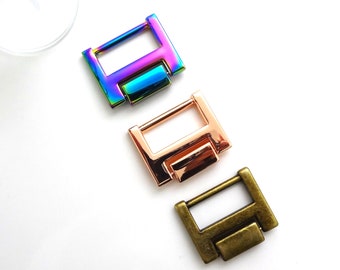 May include: Three metal slider buckles in different colors: iridescent, rose gold, and antique brass. Each buckle has a rectangular shape with a small, raised bar in the center.