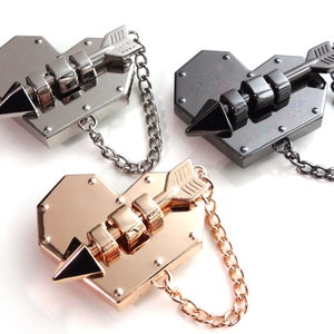 Puede incluir: Tres dijes de metal en forma de corazón con acentos de flecha y cadenas de unión. Los dijes son plateados, negros y de oro rosa.