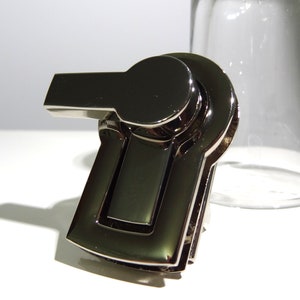 May include: A silver-toned metal clasp with a hinged latch and a rounded handle. The clasp is designed for attaching to bags, purses, or other accessories.