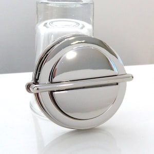 May include: A silver-toned bottle stopper with a round, flat top and a horizontal bar across the centre. The stopper is made of metal and has a polished finish.