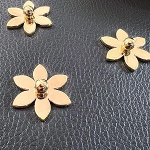 May include: Three gold metal flower-shaped decorative embellishments with a round centre. The embellishments are on a dark grey textured surface.