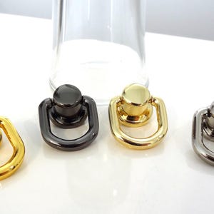 May include: Four metal ring pulls with a circular base and a hinged D-shaped loop. The set includes two polished gold-tone pieces, one dark gunmetal-tone piece, and one silver-tone piece, all featuring a smooth, reflective metallic finish.