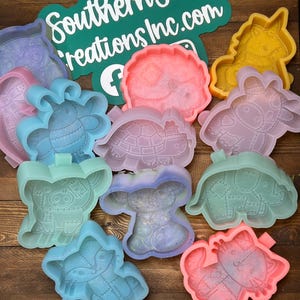May include: A collection of colorful silicone molds in various shapes, including a unicorn, turtle, and bee. The molds are in shades of pink, blue, yellow, and purple. A green sign with white text that reads "Southern Sass Creations Inc.com" is also visible.