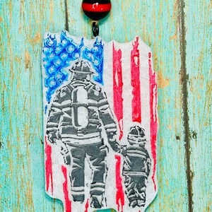 May include: A white ornament shaped like a distressed American flag with two firefighters in silhouette, one holding the hand of a smaller firefighter. The ornament has a red, white, and blue bead at the top.