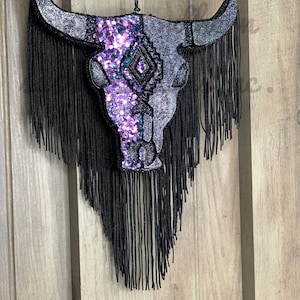May include: A black and purple sequined bull skull with black fringe hanging from a wooden wall.