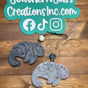 May include: Two gray manatee ornaments with blue accents and decorative swirls. Each ornament is attached to a string with a decorative bead. The background includes a teal sign with the text "Southern Sass Creations Inc.com" and social media icons.