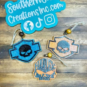 May include: Three air fresheners with skull designs. One is blue, one is orange, and one is a combination of both colors. The blue and orange fresheners have a Harley Davidson logo shape. The top of the image has a teal sign with the text "Southern Sass Creations Inc.com".