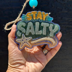 May include: A resin ornament with the words "STAY SALTY" in gold and silver lettering. The ornament has a blue border and a starfish design. It is attached to a jute string and a turquoise bead.