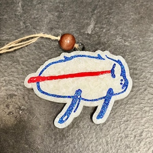 May include: A white, glittery ornament in the shape of a buffalo with blue outline and red stripe. The ornament has a brown bead and twine for hanging. The design is a sports team logo.