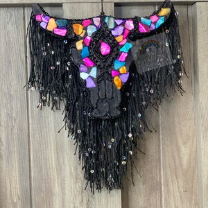 May include: A black bull skull ornament with colorful mosaic details and black fringe hanging from a wooden surface.