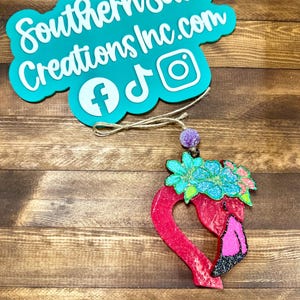 May include: Pink flamingo shaped resin ornament with a blue and green flower crown. The ornament is decorated with glitter and has a heart-shaped body. The ornament is hanging from a string with a purple bead.