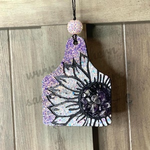 May include: Purple glitter car air freshener shaped like a cow tag with a white and black sunflower design. The center of the sunflower is filled with amethyst crystals.