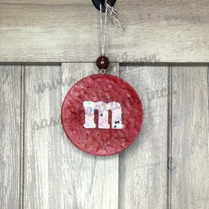 May include: Red round Christmas ornament with a white "M" in the center. The ornament is covered in glitter.
