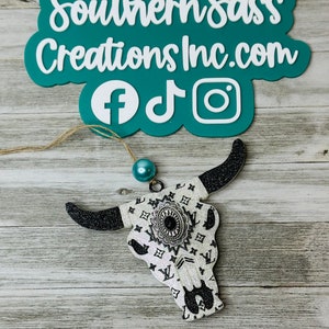 May include: White and black glitter bull skull ornament with a black and white patterned design. The ornament has a silver bead at the top and is hanging from a twine string. The ornament is on a light wood background. The text "Southern Sass Creations Inc.com" is in the background.