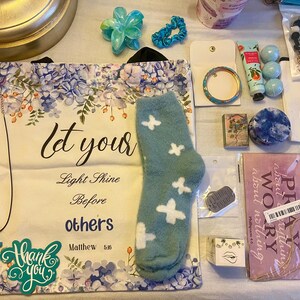 May include: An assortment of items including a tote bag with floral design and text "Let your light shine before others", a teal sock with white butterfly accents, a necklace with a cross pendant, and a bracelet.