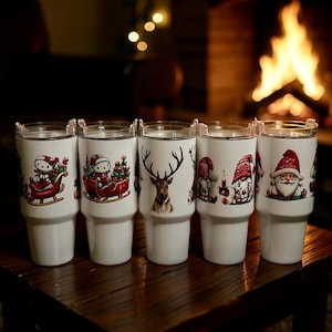 May include: Five white insulated tumblers with clear lids, each featuring a different Christmas-themed design. Designs include Santa Claus, reindeer, gnomes, and a character in a sleigh. The tumblers are arranged on a wooden surface.