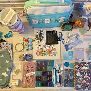 May include: An assortment of stationery and gift items, including a light blue lunch bag with "BIBLE" lettering, a tumbler, socks, journals, pens, and stickers. The items are arranged on a white surface, with floral and inspirational themes.