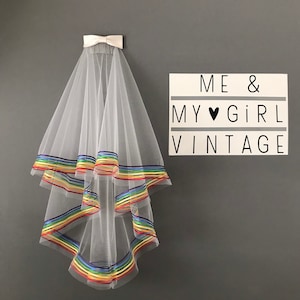 Peut inclure: Un voile de mariée en tulle blanc avec une bordure de ruban arc-en-ciel. Le voile est accroché à un mur avec une pancarte qui dit "ME & MY ♥ GIRL VINTAGE".