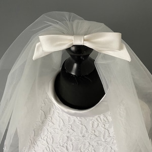 May include: A white bridal veil with a satin bow. The veil is made of sheer tulle and is attached to a black mannequin head. The dress underneath is white with a floral lace pattern.