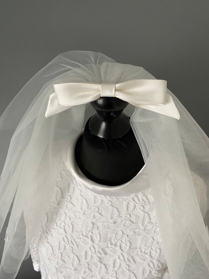 May include: A white bridal veil with a satin bow. The veil is made of sheer tulle and is attached to a black mannequin head. The dress underneath is white with a floral lace pattern.