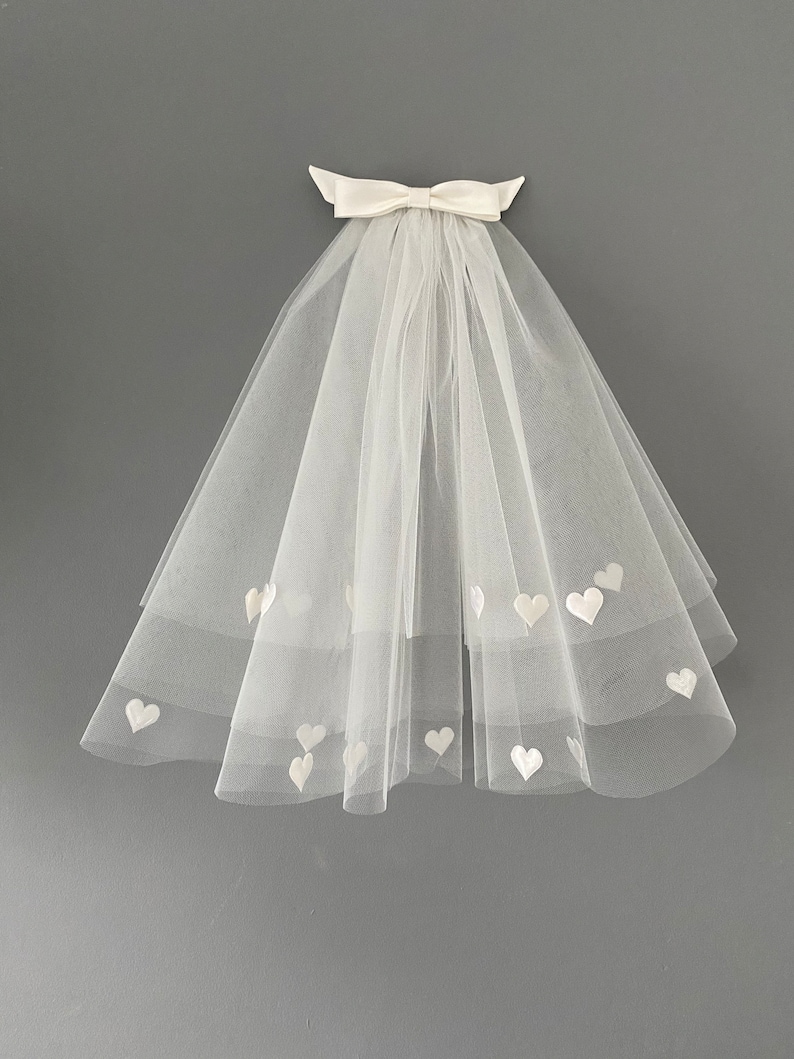 May include: A white tulle bridal veil with heart-shaped appliqu&eacute;s. The veil is adorned with a satin bow at the top. The veil is a soft, delicate accessory for a wedding or special occasion.